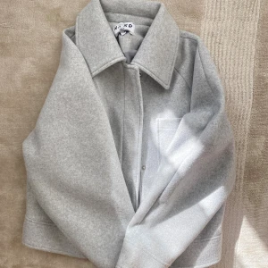 Ljusgrå overshirt från NA-KD - Snygg ljusgrå overshirt från NA-KD med klassisk krage, tryckknappar och en bröstficka. Jackan har en rak och något boxig passform, perfekt att slänga över en hoodie eller t-shirt. Materialet är mjukt och känns lite ulligt.