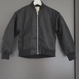 Svart bomberjacka med dragkedja - Snygg svart bomberjacka med klassisk siluett och silverfärgad dragkedja framtill. Jackan har ribbade muddar vid ärmslut och midja, samt två fickor på sidorna. Perfekt för dig som gillar en stilren och enkel look.