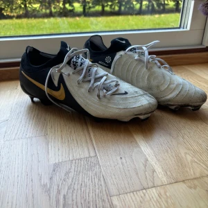 Nike fotbollsskor svart/vit/guld - Säljer ett par Nike fotbollsskor i svart, vitt och guld. Skorna har snörning, rund tå och platt sula med dobbar för bra grepp på planen. Snyggt guldigt swoosh-märke på sidan och stickad detalj vid ankeln. Perfekta för dig som vill sticka ut på matchen.