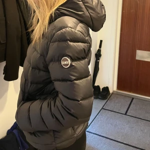 Svart pufferjacka från Colmar - Säljer en svart pufferjacka från Colmar Originals med huva och dragkedja framtill. Jackan har ett stilrent, quiltat mönster och logotyp på ärmen. Perfekt för kalla dagar och har en snygg, normal passform som funkar till de flesta outfits.