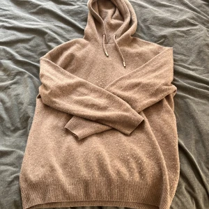 Beige stickad hoodie med huva - Mysig beige stickad hoodie med huva och snörning. Tröjan har långa ärmar och ribbade muddar vid ärmslut och nederkant. Perfekt för kyliga dagar när du vill vara både bekväm och snygg.