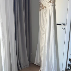 Bröllopsklänning med spets och slits - Superfin oanvänd ny bröllopsklänning med transparent spets upptill med blommiga detaljer. Klänningen har en snygg slits framtill och ett omlottband kommer med satin band för ryggen. Finns att hämta i Vasastan även prövning är möjligt.