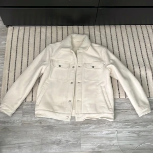 Vit teddyjacka med fickor - Säljer en vit teddyjacka med mjuk krage och två stora bröstfickor med knappar. Jackan har knappstängning framtill och är långärmad. Perfekt för dig som gillar stilren och clean look med lite extra fluff vid halsen.