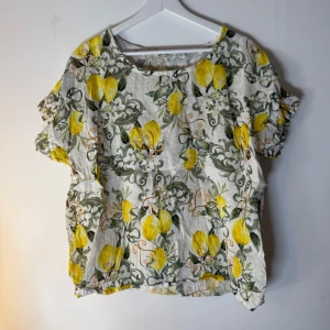 Mönstrad blus  - Vit blus med ett tryck av citroner, gröna blad och vita blommor. Blusen har korta ärmar med volang. Storlek L. Gjord av 100% linne 