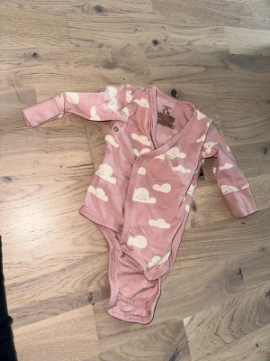 Rosa omlottbody med moln från Lindex Rosa Klänning och Body -  Lindex - Supersöt rosa omlottbody från Lindex i storlek 56. Bodyn har ett gulligt mönster med vita moln med små ansikten och praktiska tryckknappar längs sidan och i grenen. Tillverkad i mjuk bomull som är skön mot huden.Ett sött rosa klänning med broderade detaljer och volangärmar. En rosa omlottbody med moln tryck. 2-4 mån