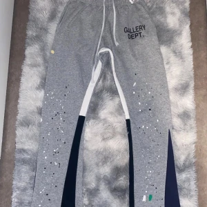 Grå mjukisbyxor Gallery Dept. - Grå mjukisbyxor från Gallery Dept. med vit snörning i midjan och tryckt logga på vänster höft. Byxorna har färgstänk i vitt, svart, grönt och gult samt mörkblå paneler längs benen. Tillverkade i 100% bomull för en skön och avslappnad vibe.