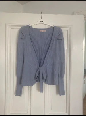 Ljusblå ribbad kofta Soft Rebels - Säljer en ljusblå ribbad kofta från Soft Rebels i storlek S. Koftan har långärmade ärmar, omlottmodell med knytband framtill och är stickad i mjukt material. Perfekt för dig som gillar chill och stilren look.