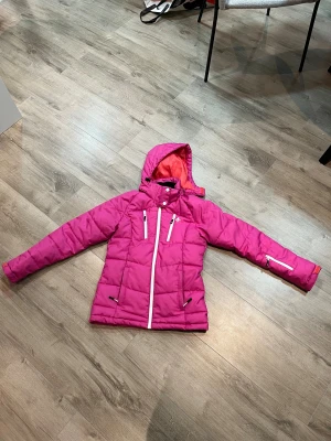 Rosa pufferjacka från Cross Sportswear XS - Snygg rosa pufferjacka från Cross Sportswear i storlek XS. Jackan har huva, vita dragkedjor och flera fickor framtill. Insidan är korallröd och materialet är syntet. Perfekt för kalla dagar med sin quiltade design och långa ärmar.