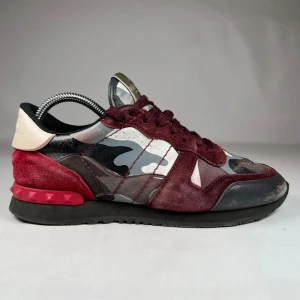 Valentino Rockrunner sneakers vinröd - Snygga Valentino Rockrunner sneakers med vinröda mockadetaljer, svart och grått kamouflagemönster samt röda nitar på hälen. Skorna har snörning, platt sula och rund tå. Materialmix av mocka, skinn och textil ger en lyxig känsla. Perfekt för dig som vill sticka ut med coola detaljer.