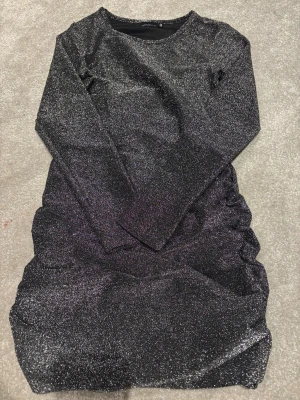 Glittrig svart klänning XOXO Friends - Supersnygg svart glittrig klänning från XOXO Friends i kort modell. Klänningen har lång ärm och är gjord i ett stretchigt syntetmaterial som glittrar i ljuset. Perfekt till speciella högtider, nyår eller fest.