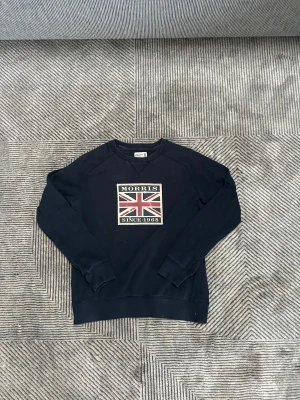 Mörkblå sweatshirt Morris  - Snygg mörkblå sweatshirt från Morris i storlek S. Framsidan har ett stort Union Jack-tryck med texten 'Morris Since 1968'. Tröjan har rund hals, långa ärmar och ribbade muddar. Perfekt för dig som gillar klassisk stil med en brittisk touch.