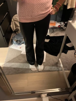 Svarta bot cut byxor i skinnimitation - Säljer ett par svarta byxor i skinnimitation med bot cut passform. Byxorna har en snygg, slät yta och sitter tajt över höften och låren, sedan faller de rakt ner mot ankeln. Perfekt för dig som vill ha en cool och stockolmstil look, använd fåtal gånger