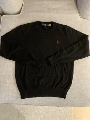 Svart stickad tröja från Polo Ralph Lauren - Svart stickad tröja från Polo Ralph Lauren med klassisk rund hals och långärmad passform. Tröjan har en röd broderad logga på bröstet och ribbade muddar vid ärmslut och nederkant. Perfekt för dig som gillar stilrena och tidlösa plagg.