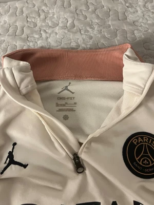 PSG x Jordan långärmad träningströja - Snygg långärmad träningströja från PSG och Jordan med halv dragkedja, beige färg och kontrasterande ljusrosa krage. Tröjan har tryck med Qatar Airways och Paris på ärmen, samt PSG-logga och Jumpman på bröstet. Perfekt för fotbollsträning eller chill.