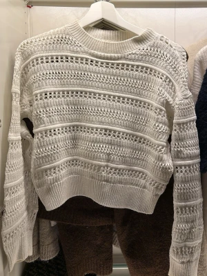 Vit stickad tröja från H&M - Säljer en vit stickad tröja från H&M med snyggt hålmönster och ribbade muddar. Tröjan har lång ärm och rund halsringning, perfekt för lager-på-lager och chill stil. Passar dig som gillar en fräsch och enkel look.