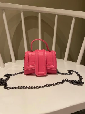 Mini rosa axelväska med kedja - Superfin liten axelväska i rosa med croco-mönster och silverfärgad kedja. Väskan har ett topphandtag och en unik frontdetalj som sticker ut. Perfekt för dig som vill ha en färgklick till outfiten.