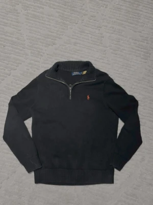 Svart stickad tröja Polo Ralph Lauren quarter zip - Svart stickad tröja från Polo Ralph Lauren med röd broderad logga på bröstet. Tröjan har ribbad krage, muddar och nederkant samt en half zip-dragkedja vid halsen. Perfekt för dig som gillar klassisk och stilren design.