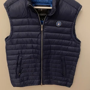 Marinblå dunväst från Sail Racing - Snygg marinblå dunväst från Sail Racing med quiltad design och två dragkedjeförsedda fickor. Västen har hög krage, logomärke på bröstet och hel dragkedja framtill. Perfekt för lager-på-lager och stilren look.