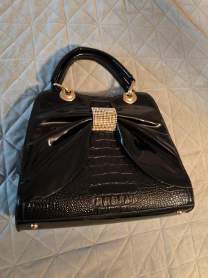 Assessorer  - Snygg svart handväska – i nyskick! Säljer en elegant och rymlig handväska i glansigt svart material. Väskan är perfekt för både vardag och fest, och rymmer utan problem allt du behöver – mobil, plånbok, smink, småsaker och mer. Den är lätt att bära och har en stilren design som passar till alla outfits. Väskan är i mycket fint skick, nästan som ny. Den är endast använd 2–3 gånger, så den ser fortfarande väldigt fräsch ut.