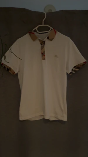 Vit Burberry piké med rutig krage - Säljer en vit Burberry t-shirt med klassisk rutig krage och detaljer vid ärmslut. T-shirten har korta ärmar och en liten broderad logga på bröstet. Materialet är mjuk bomull och passformen är normal. Perfekt för dig som gillar stilrena designer med lyxig touch.