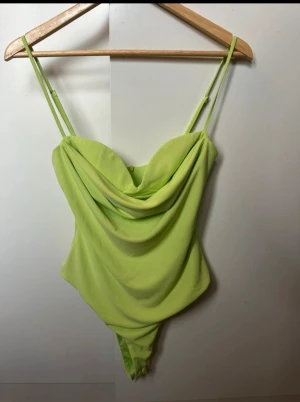 Ljusgrön omlott bodysuit från Zara - Säljer en ljusgrön bodysuit från Zara med omlott-drapering framtill och smala axelband. Toppen har en snygg, croppad passform och är tillverkad i mjukt syntetmaterial. Perfekt för dig som vill sticka ut med en cool färg och trendig look.