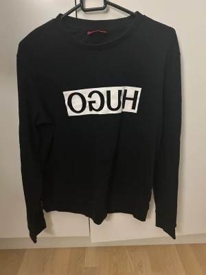 Svart långärmad tröja från Hugo - Svart långärmad tröja från Hugo med vit logotyp tryckt i spegelvänd stil på bröstet. Tröjan har rund hals och är tillverkad i mjuk bomull. Snygg och enkel design med en modern vibe, perfekt för dig som gillar streetwear och tydliga loggor.