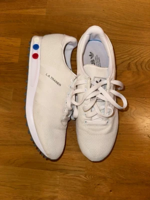 Adidas LA Trainer vita sneakers strl 42 - Säljer ett par Adidas LA Trainer sneakers i vit färg med mesh-ovandel och klassisk snörning. Skorna har en platt sula med gummidubbar och detaljer i rött och blått på sidan av sulan. Adidas-logga på plös och innersula. Perfekt för dig som gillar retrostil. Storlek 42