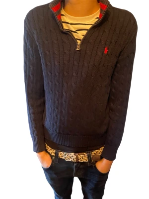 Marinblå stickad half zip från Polo Ralph Lauren - Kabelstickad marinblå tröja från Polo Ralph Lauren med röd broderad logga på bröstet. Tröjan har half zip-dragkedja, ribbade muddar och hög krage med röd detalj på insidan. Perfekt för dig som gillar klassisk och stilren look. Priset är inte hugget i sten