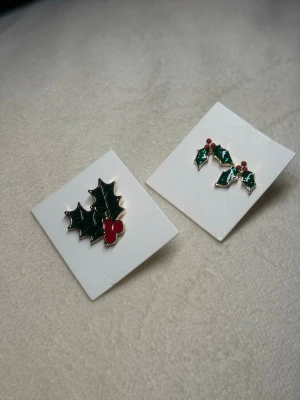 Juligt Smyckeset med Järnek – Brosch & Örhängen – Perfekt Julklapp! - Ett charmigt och riktigt festligt smyckeset med klassiskt järnek/julblad-motiv, perfekt för att skapa en mysig och lekfull julkänsla. Setet består av: 	•	En dekorativ brosch med gröna blad och röda bär 	•	Matchande örhängen i samma juliga design Båda delarna är helt nya och sitter kvar på sina kort. De klara färgerna och den glansiga emaljen gör setet till ett riktigt fint blickfång under hela december – perfekt till jultröjan, festklänningen eller som en söt detalj på ytterplagget. 
