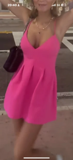 Rosa kort klänning med v-ringning - Supersöt rosa kort klänning med v-ringad hals och smala axelband. Kjolen har lätt utställd form med veck som ger extra volym. Perfekt för dig som vill sticka ut med en färgstark look.