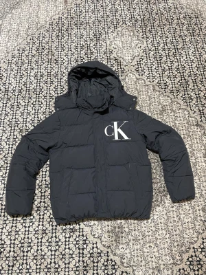 Svart pufferjacka Calvin Klein Jeans - Svart pufferjacka från Zalando med stor vit CK-logga på bröstet. Jackan har huva, långa ärmar och två fickor framtill. Materialet är syntet och jackan är quiltad för extra värme. Perfekt för kalla dagar och har en clean modern look. Säljer den eftersom den passar inte, den är storlek M. Den är använd bara några gånger. Jag tror jag har fortfarande kvittot kvar. Skriv privat om du är intresserade!!! Pris kan diskuteras!✅
