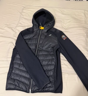 Svart pufferjacka Parajumpers S - Svart pufferjacka från Parajumpers med huva, quiltad framsida och coolt PJS-märke på ärmen. Jackan har dragkedja, bröstficka med zip och snygg gul detalj vid halsen.Perfekt för kyliga dagar.