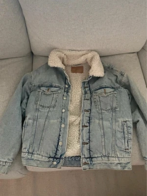 Ljusblå jeansjacka med teddyfoder - Säljer en ljusblå jeansjacka från Denim med mjukt vitt teddyfoder på insidan och kragen. Jackan har klassiska bröstfickor med knapp, sidofickor och silverfärgade knappar framtill. Perfekt för dig som vill ha en chill och varm look. Den är nog lite oversize för jag är M och den sitter på mig som man kan se bild nr 5. 