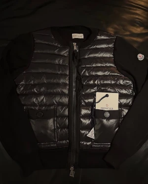 Moncler Cardigan  - Snygg svart pufferjacka från Moncler med quiltad framsida och stickade ärmar. Jackan har rund hals, två fickor med knappstängning och dragkedja framtill. Moncler-logga på ärmen och coola detaljer som ger en exklusiv vibe.