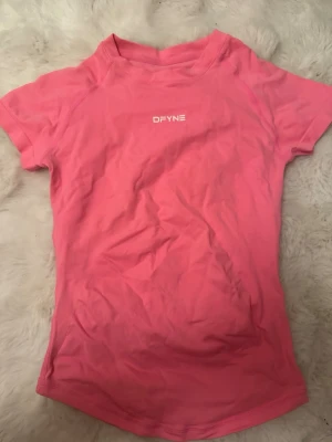 Rosa träningstopp från DFYNE - Säljer en snygg rosa träningstopp från DFYNE i storlek small. Toppen har korta ärmar, rund hals och är gjord i ett stretchigt material med 90% nylon och 10% elastan. Perfekt för gymmet eller sport, med logga framtill och skön passform.