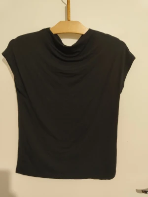 Svart top. - Svart topp med snygg draperad halsringning och ärmlös design. Enkel och stilren form som passar till många olika looks. Materialet är mjukt och skönt.