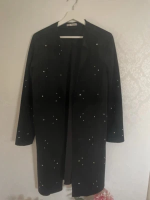 Svart rock/kappa med pärldetaljer / Nitar från Vila - Snygg svart kappa eller rock från Vila med pärldetaljer över hela jackan. Rak och längre modell med rundad hals och lång ärm. Perfekt för dig som gillar stilrena plagg med lite extra bling. Materialet känns som mocca. Faux 