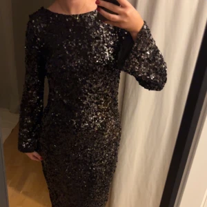 Svart glittrig långklänning med öppen rygg - Svart långklänning med glittriga paljetter över hela klänningen. Modellen har lång ärm, tight passform och en djup öppen rygg med knytband i nacken. Perfekt klänning för nyår✨prislapp kvar! Är till fötterna på mig som är 175 cm lång.