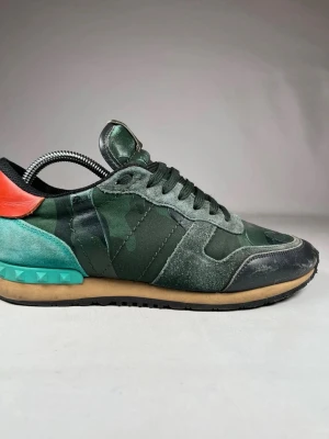 Valentino Garavani Rockrunner sneakers - Snygga Valentino Garavani Rockrunner sneakers med camouflagemönster i olika gröna toner, svart och turkos detalj på hälen samt röd accent. Skorna har snörning, platt sula och är tillverkade i en mix av mocka, skinn och textil. Perfekt för dig som gillar statement-sneakers. Skriv om ni har frågor eller funderingar!😊