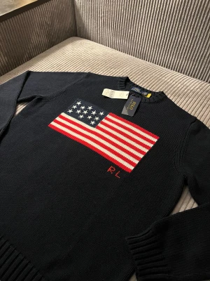 Mörkblå stickad tröja Polo Ralph Lauren - USA - Snygg mörkblå stickad tröja från Polo Ralph Lauren med amerikansk flagga i rött, vitt och blått på bröstet samt RL-broderi. Tröjan har rund hals, ribbade muddar och lång ärm. Perfekt för dig som gillar klassisk och cool stil. Längd: 67,5 cm