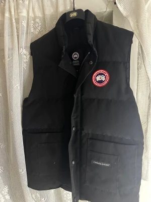 Svart dunväst från Canada Goose - Svart dunväst från Canada Goose med klassisk logga på bröstet. Västen har hög krage, två stora fickor framtill och stängs med både dragkedja och tryckknappar. Perfekt för lager-på-lager och har en clean, stilren look.