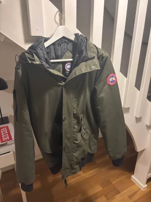 Grön vinterjacka från Canada Goose - Säljer en grön vinterjacka från Canada Goose med huva och svarta muddar. Jackan har dragkedja och knappar framtill, samt fickor med lock och ärmficka med knapp. Klassisk Canada Goose-logga på ärmen och broderad logotyp på ryggen.