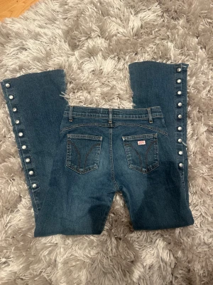 Blå bootcut jeans med knappar Miss Sixty - Snygga blå jeans från Miss Sixty med bootcut passform och coola silverfärgade knappar längs hela utsidan av benen. Jeansen har knappgylf, klassiska fickor och är tillverkade i mjukt jeansmaterial. Perfekt för dig som gillar detaljer och unik stil.
