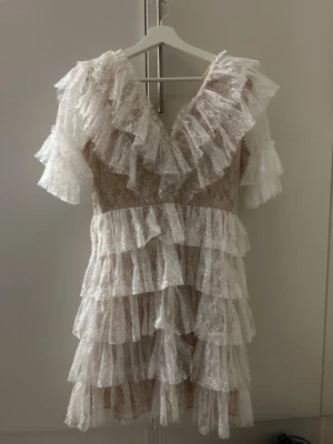 Vit spetsklänning med volanger - Superfin kort klänning i vit spets med flera lager volanger och v-ringad hals. Klänningen har korta ärmar och är tillverkad i ett tunt, romantiskt spetsmaterial som ger en drömmig känsla. Perfekt för dig som gillar detaljer och en feminin look. Andvänd endast en gång.