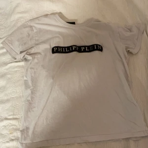 Vit Philipp Plein t-shirt med tryck - Säljer en vit t-shirt från Philipp Plein med svart tryck över bröstet. Modellen är klassisk med rund hals och korta ärmar. Materialet är mjuk bomull och passformen är normal. Perfekt för dig som gillar exklusiva märken och stilren design.