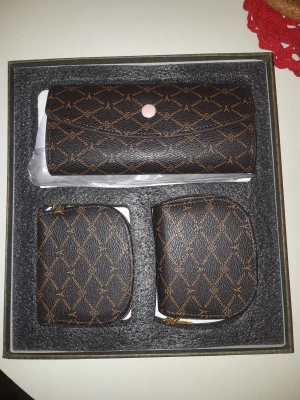 Set med bruna plånböcker med mönster - Snyggt set med tre bruna plånböcker från Amodi, alla med ett elegant rutigt mönster och guldfärgade dragkedjor. Den största har en rosa knapp och passar perfekt för kort och kontanter. De mindre är smidiga för mynt och småsaker. Perfekt för dig som gillar stilrena accessoarer.