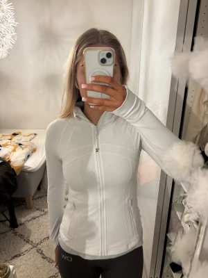 Vit zip-hoodie från Lululemon - Snygg vit hoodie från Lululemon med dragkedja hela vägen, huva och två fickor med zip. Figurnära passform och coola sömdetaljer. Materialet är mjukt och stretchigt med 87% nylon och 13% elastan, perfekt för träning eller chill.