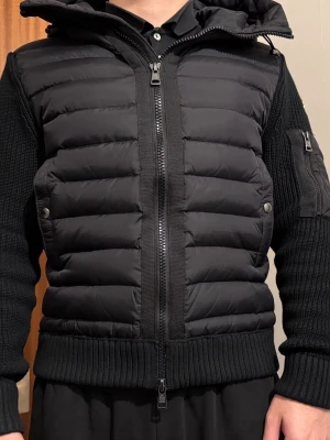 Svart Moncler cardigan med stickade ärmar - Svart pufferjacka från Moncler med stickade ärmar och ribbad mudd. Jackan har tvåvägsdragkedja framtill, fickor med tryckknappar och en extra ficka med dragkedja på ärmen. Moncler-logga på vänster ärm. Perfekt för dig som gillar exklusiv streetwear.