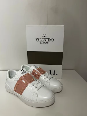 Valentino sneakers - Snygga vita Valentino sneakers med ljusrosa band och ikoniska pyramidnitar. Skorna har rund tå, platt sula och snörning. Tillverkade i lyxigt skinn med coola detaljer som gör dem till ett statement. Perfekta för dig som gillar exklusiv streetstyle.