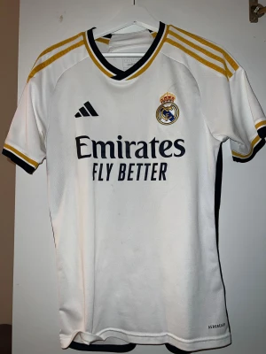 Real Madrid Bellingham tröja S - Säljer en Real Madrid matchtröja från Adidas i storlek S med Bellingham och nummer 5 på ryggen. Tröjan är vit med svarta och gula detaljer, har broderat klubbmärke och Adidas-logga samt 'Emirates Fly Better' tryck på bröstet. Slim fit och tillverkad i polyester. Köpt för 1499kr äkta.